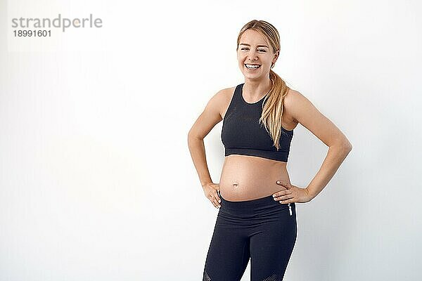 Glückliche  gesunde  junge  schwangere Frau in schwarzer Sportkleidung  die mit den Händen auf den Hüften steht und mit einem strahlenden Lächeln in die Kamera schaut