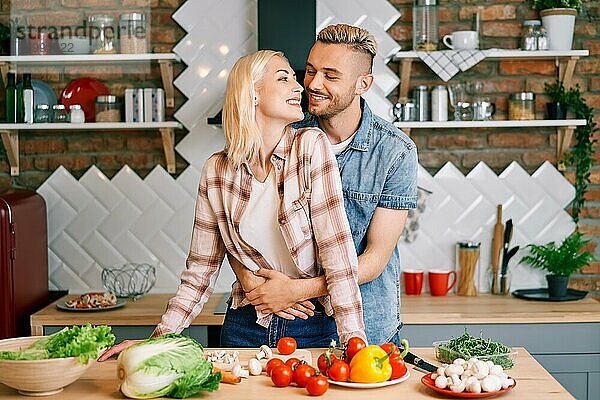 Lächelndes junges Paar kocht zusammen vegetarische Mahlzeit in der Küche zu Hause. Mann umarmt Frau. Gesunder Lebensstil Konzept