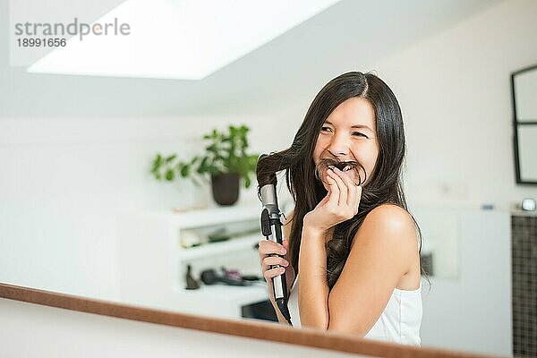 Verspielte junge Frau  die sich die Haare stylt und mit einer ihrer langen Strähnen einen Schnurrbart vortäuscht  während sie im Badezimmerspiegel vor sich hin grinst