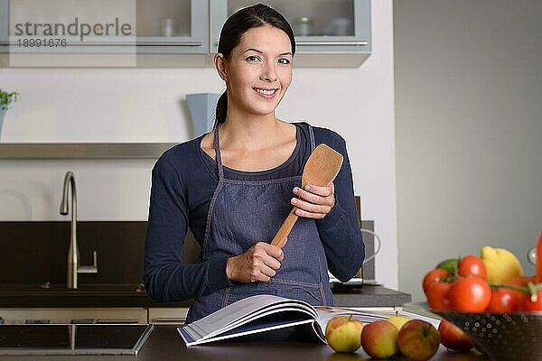 Glückliche hübsche Frau mit Schürze  die eine hölzerne Schöpfkelle hält  während sie ein Rezeptbuch am Tisch mit Obst und Gemüse in der Küche liest