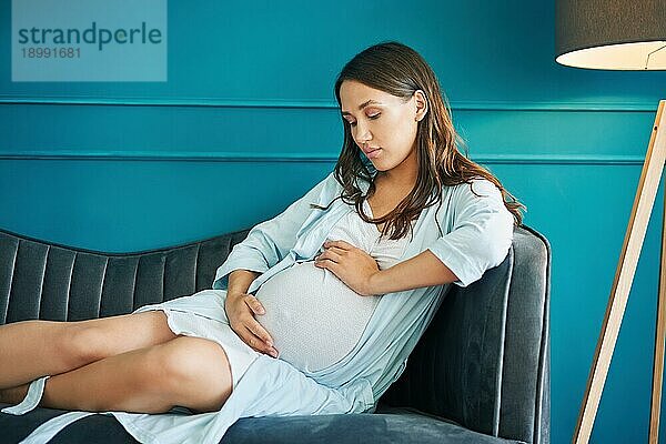 Porträt der jungen schwangeren Frau  die ihren Bauch berührt  entspannt auf dem Sofa in einem modernen Haus. Schwangerschaft  Mutterschaft  Menschen und Erwartung Konzept