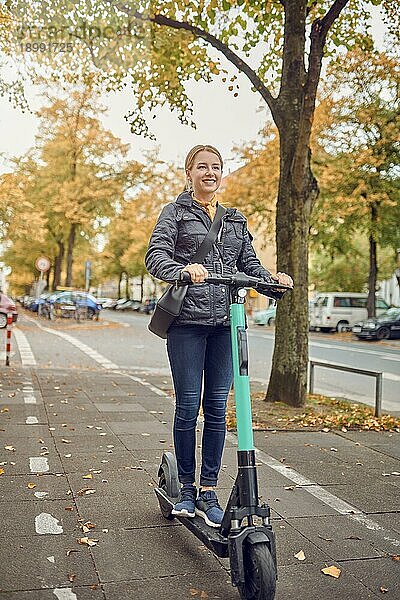 Junge glückliche blonde Frau fährt mit einem Elektroroller in der Stadt  lächelt in die Kamera  im Herbst