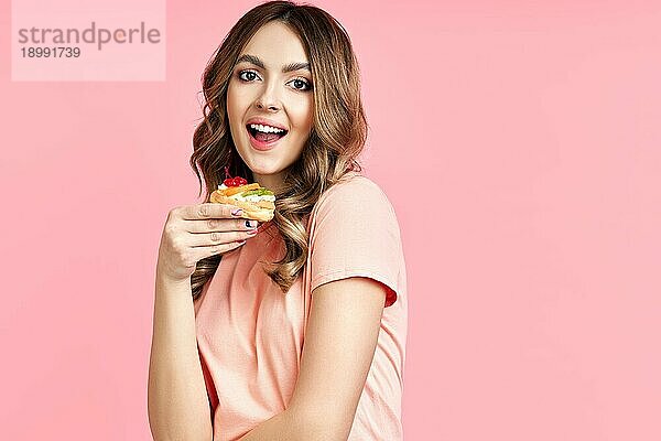 Schöne lächelnde junge Frau mit einem leckeren Gebäck Kuchen auf rosa Hintergrund. Lebensmittelkonzept