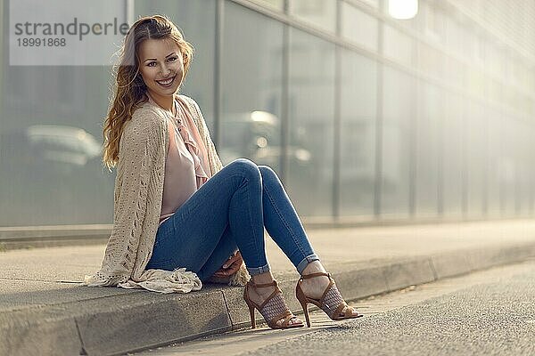 Lächelnde  hübsche  trendige junge Frau  die sich auf dem Bürgersteig einer städtischen Straße in ihren hochhackigen Schuhen und ihrem modischen Outfit ausruht und in die Kamera lächelt