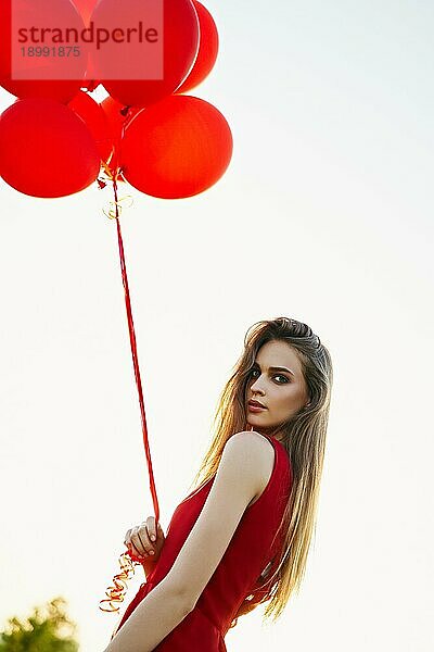 Junge schöne Frau in rotem Kleid posiert im grünen Feld mit roten Luftballons bei Sonnenuntergang. Freiheit  Spaß  Mode Konzept