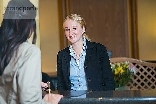Schöne stilvolle blonde Hotelrezeptionistin steht hinter dem Service Schreibtisch in einer Hotellobby und sieht einen Hotelgast mit einem freundlichen Lächeln an