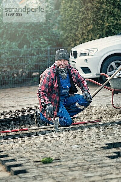 Fröhlicher Arbeiter in warmer Winterkleidung kniet am Straßenrand und verlegt Pflastersteine in seiner Latzhose und schaut mit einem freundlichen Lächeln in die Kamera