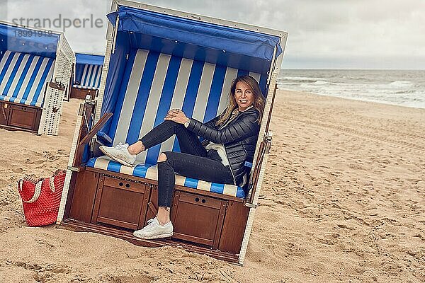 Attraktive schlanke blonde Frau  die in einer Strandhütte mit blauweiß gestreiftem Stoff an einem Sandstrand an einem bewölkten Tag sitzt und in die Kamera lächelt