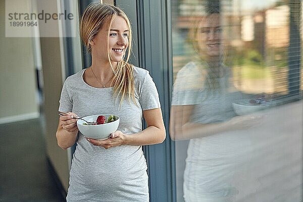 Glückliche  gesunde  schwangere  junge  blonde Frau  die an ein Terrassenfenster gelehnt steht und lächelnd nach draußen schaut  während sie eine Schüssel mit frischem Obstsalat genießt