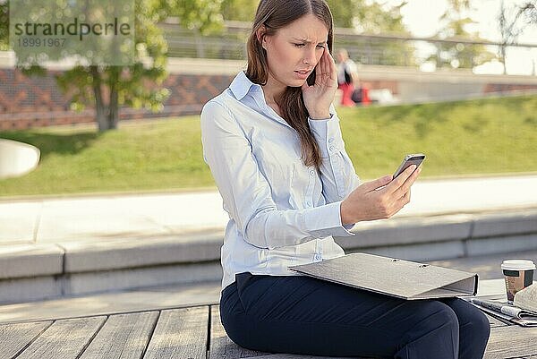 Attraktive schlanke junge Frau  die im Freien in einem Stadtpark sitzt und mit besorgtem Blick und der Hand im Gesicht eine SMS auf ihrem Handy liest