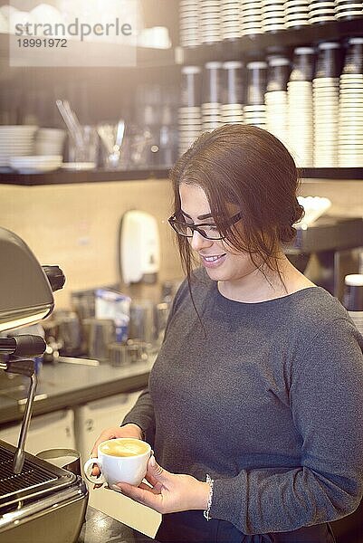 Weibliche Barista oder Kellnerin hält eine Tasse Cappuccino in einem Kaffeehaus oder einer Cafeteria  lächelnd und zufrieden