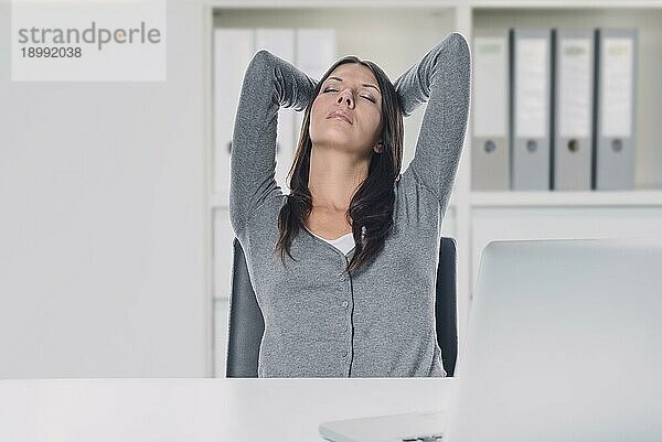 Junge Geschäftsfrau  die einen entspannenden Moment genießt  während sie in ihrem Stuhl am Schreibtisch sitzt  die Hände hinter dem Kopf verschränkt und die Augen mit einem heiteren Ausdruck geschlossen hat