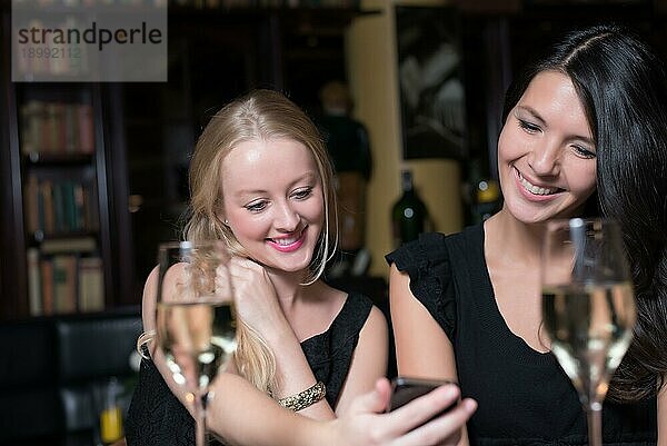 Zwei schöne junge Frauen genießen ein Glas Wein in einer Nacht mit ihren Handys und lachen über das Gespräch und die Textnachricht
