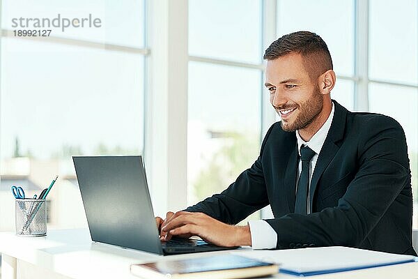 gutaussehender lächelnder Geschäftsmann im eleganten Anzug  der in einem hellen modernen Büro am Laptop arbeitet. geschäftliches Erfolgskonzept