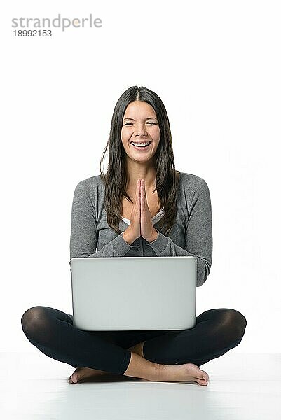 Glückliche junge Frau meditiert mit ihrem Laptop im Schneidersitz und barfuß auf dem Boden sitzend  die Hände wie zum Gebet gefaltet und mit einem strahlenden Lächeln der Freude  vor weißem Hintergrund