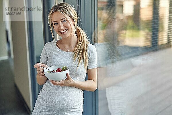 Glückliche  gesunde  schwangere  junge  blonde Frau  die an ein Terrassenfenster gelehnt steht und in die Kamera lächelt  während sie eine Schüssel mit frischem Obstsalat genießt