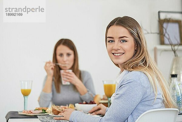 Hübsche junge Frau im Teenageralter mit lebhaftem Lächeln  die mit ihrer besten Freundin an einem Tisch sitzt und das Frühstück genießt und sich zur Kamera dreht