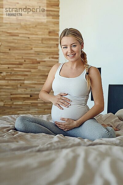 Glückliche schwangere Frau sitzt auf einem Bett und wiegt ihren Babybauch mit ihren Händen und lächelt in die Kamera
