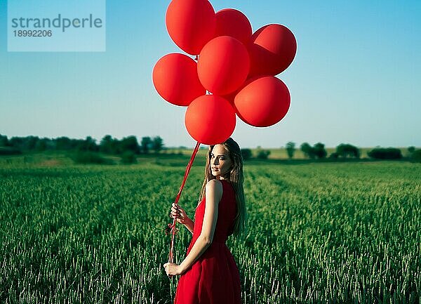 Junge schöne Frau in rotem Kleid posiert im grünen Feld mit roten Luftballons. Freiheit  Spaß  Urlaub Konzept