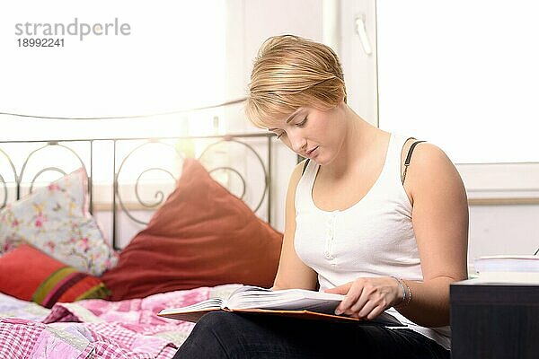 Hübsche junge Studentin  die in einem Lehrbuch liest  während sie in ihrem Zimmer sitzt und die Sonne durch das Fenster scheint