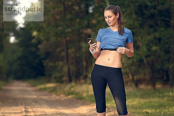Junge  sportliche Frau  die im Wald trainiert und auf ihr Smartphone schaut  während sie eine mobile Anwendung nutzt  die elektronisch mit ihrem Pulsgurt verbunden ist