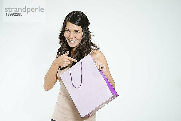 Glückliche Einkäuferin  die mit einem spielerischen  verschwörerischen Blick auf ihre Einkaufstasche zeigt  während sie zufrieden über den Kauf eines Überraschungsgeschenks lächelt  isoliert mit Kopierraum