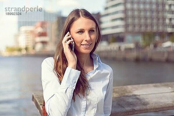 Attraktive Frau  die einen Anruf mit ihrem Mobiltelefon entgegennimmt  steht im Freien vor einem Flusshintergrund und lächelt in die Kamera  während sie dem Gespräch zuhört