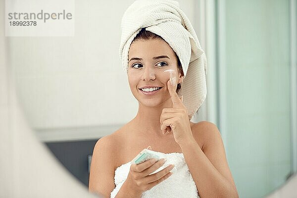 Hübsche junge Frau in weißen Handtüchern in einem Badezimmer  die der Kamera ein freundliches Lächeln schenkt  während sie ihre Wangen eincremt und ein Hautpflege und Hygienekonzept verfolgt