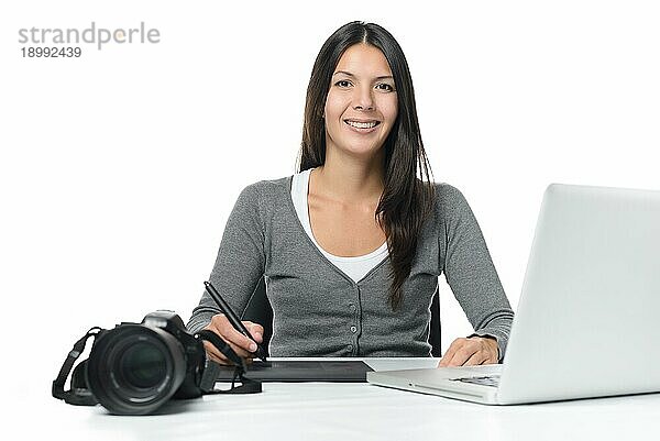 Weibliche Fotografin lächelt  während sie ihre kürzlich heruntergeladenen Dateien auf einem Laptop mit einem Tablet und einem Stift bearbeitet  während ihre Kamera daneben auf dem Schreibtisch liegt  über weiß