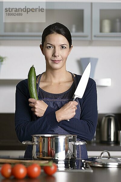 Glückliche junge Frau posiert in ihrer Küche mit verschränkten Armen  ein Messer und frische Gurken haltend  während sie ein gesundes Abendessen zubereitet  Frontalansicht am Herd stehend