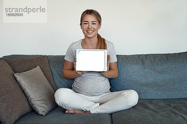 Lächelnde  attraktive  lebhafte  junge  schwangere Frau  die im Schneidersitz auf einem Sofa zu Hause sitzt und ein Tablet mit leerem Bildschirm und Kopierbereich hochhält