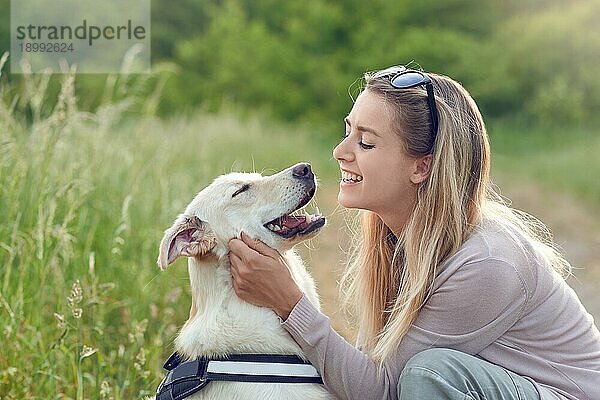 Glücklich lächelnder goldener Hund  der ein Laufgeschirr trägt und seiner hübschen jungen Besitzerin gegenübersitzt  die ihn mit einem liebevollen Lächeln im Freien auf dem Lande streichelt