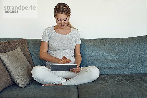 Glückliche schwangere Frau  die sich auf einem Sofa im Schneidersitz entspannt und ein eBook auf einem Tablet liest  mit einem warmen  freundlichen Lächeln