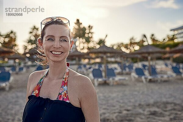 Attraktive glückliche stilvolle Frau am Strand bei Sonnenuntergang mit Liegestühlen hinter ihr auf dem Sand schaut zur Seite mit einem schönen lebhaften Lächeln an einem Sommerabend