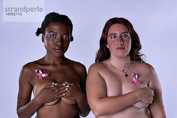 Studioporträt von zwei jungen Frauen  die ihre Brüste bedecken und in die Kamera schauen  mit einer rosa Schleife an der Brust. Brustkrebs Bewusstsein Konzept