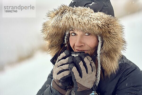 Glücklich lächelnde Frau in einer pelzbesetzten Kapuzenjacke  die eine Tasse Kaffee in ihren behandschuhten Händen im Freien im Winterschnee in einem Konzept der Jahreszeiten in einer Nahaufnahme genießt