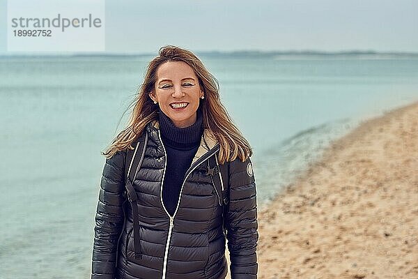 Attraktive Frau  die an einem bewölkten Tag am Strand spazieren geht  einen Rucksack und eine Jacke trägt und am Rande des Meeres innehält  um in die Kamera zu lächeln