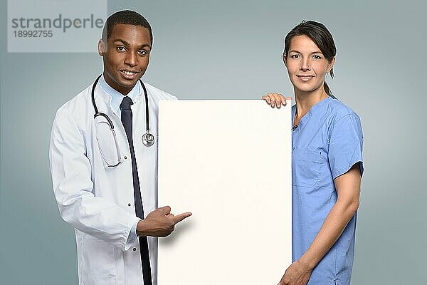 Multiethnisches Ärzteteam mit einem afrikanischen Arzt im Laborkittel und Stethoskop und einer Krankenschwester in OP Kleidung  die ein leeres weißes Schild halten  auf dem Ihr Text mit einem freundlichen Lächeln zu lesen ist