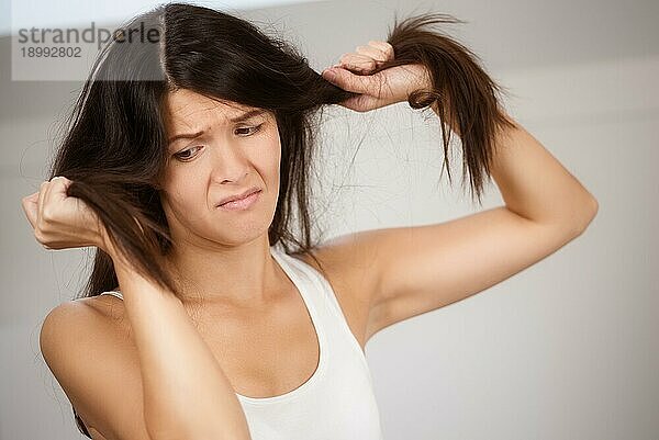 Attraktive junge Frau  die mit dem Zustand ihres langen Haars unzufrieden ist  steht grimassierend vor der Kamera und hält mit den Händen  die sie in ihren Locken verschränkt hat  die Haare auf beiden Seiten ihres Gesichts heraus