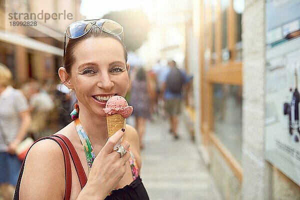 Lächelnde attraktive stilvolle Frau mit Sonnenbrille auf der Stirn  die ein Eis in einer Waffel im Freien in einer Fußgängerzone in einer Stadt genießt