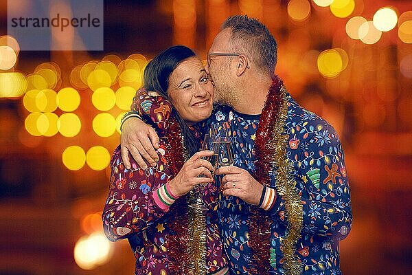 Joviales romantisches Paar mittleren Alters  das sich küsst und Champagner trinkt  um die Weihnachtszeit zu feiern  gekleidet in farbenfrohe weihnachtliche Oberteile vor einem Bokeh aus funkelnden Partylichtern