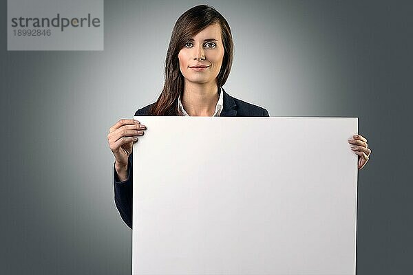 Attraktive Geschäftsfrau  die ein leeres weißes Schild vor ihrer Brust mit Platz für Ihren Text oder Ihre Werbung vor einem grauen Hintergrund in den Händen hält