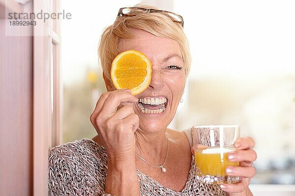 Lachende  lebensfrohe blonde Frau mittleren Alters  die sich eine Orangenscheibe ans Auge hält  während sie in der anderen Hand ein Glas frisch gepressten Orangensaft hält  in einem gesunden Ernährungskonzept