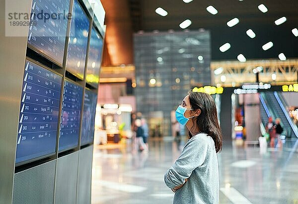 Frau mit Gesichtsmaske zum Schutz vor Viren schaut auf eine Informationstafel  um ihren Flug auf einem internationalen Flughafen zu überprüfen. Abflugtafel  Flugstatus