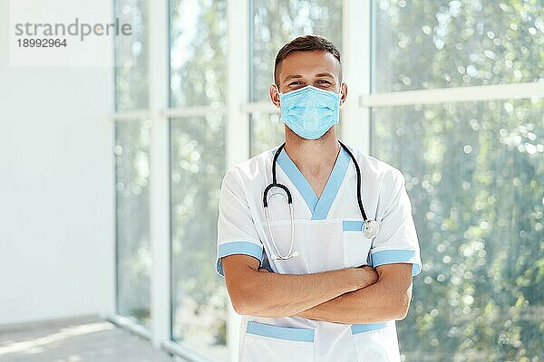 Selbstbewusster männlicher Arzt mit medizinischer Maske und verschränkten Armen im Krankenhaushintergrund. Medizin  Beruf und Gesundheitswesen Konzept