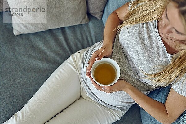 Draufsicht auf eine junge schwangere Frau  die sich zu Hause auf einem bequemen Sofa mit einer großen Tasse heißen Tees zurücklehnt  in einer Nahaufnahme auf ihren Bauch