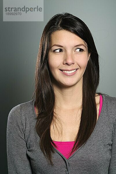 Attraktive junge Frau mit schönem  freundlichem Lächeln  die mit abgewandtem Blick seitlich nach links blickt  Kopf Schulter Studioporträt auf Grau