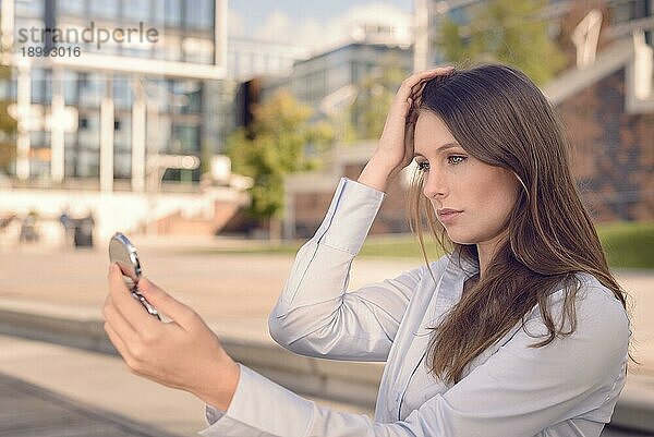 Schöne junge Frau  die ihr Aussehen in einem kleinen Handspiegel auf der Straße überprüft und lächelt  während sie ihre langen braunen Haare aus dem Gesicht hält