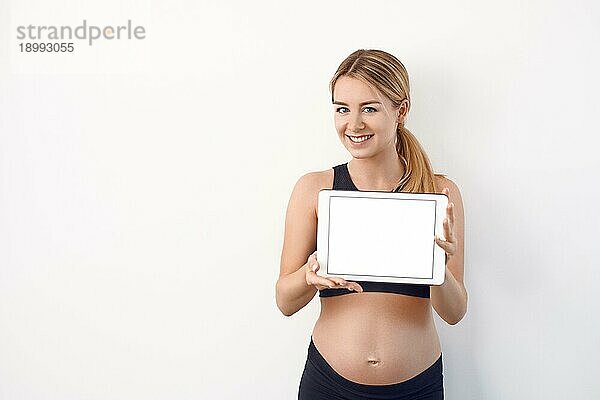 Lächelnde freundliche junge schwangere Frau  die dem Betrachter ein weißes modernes Tablet mit Bildschirm vor einem Studiohintergrund mit Kopierraum entgegenhält