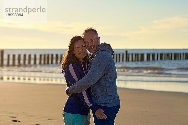 Glückliches romantisches Paar mittleren Alters genießt eine liebevolle Umarmung an einem verlassenen tropischen Strand bei Sonnenuntergang und lächelt in die Kamera mit Kopie Raum